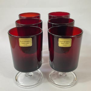 Könnte beinhalten: Satz mit sechs roten Glasbechern mit klaren Glasstielen und -böden. Jeder Becher hat ein goldenes Etikett mit der Aufschrift "Luminarc Verre d'Art France".