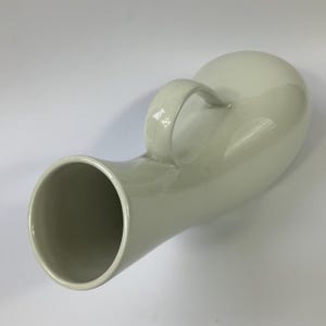 以下が含まれることがあります： クリーム色のセラミック製小便器、取っ手付き。 小便器は片側に丸みを帯びた球根状の形をしており、もう一方には円筒形の開口部があります。 ハンドルはアーチ型で、小便器の本体に取り付けられています。