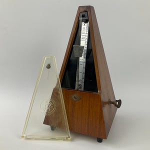 Könnte beinhalten: Ein hölzernes Metronom mit einer transparenten Kunststoffabdeckung. Das Metronom hat eine Pyramidenform mit einem Pendel und einer Skala. Die transparente Kunststoffabdeckung hat ein kreisförmiges Design. Das Metronom wird verwendet, um die Zeit in der Musik einzuhalten.