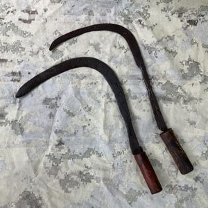 May include: Two antique, curved, metal sickles with wooden handles. The sickles are black and have intricate designs on the blades. The handles are brown and have a worn look.