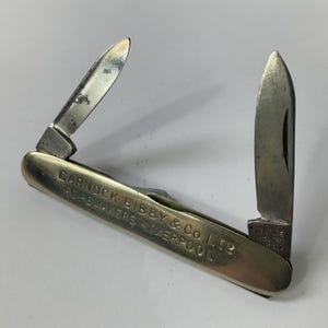 May include: A vintage pocket knife with two open blades. The knife has a silver-colored metal handle with the text "GARNOCK BIBBY & Co LTD ROPE MAKERS LIVERPOOL" engraved on it. The blades show signs of wear.