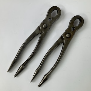 Puede incluir: Dos cascanueces antiguos de metal con un acabado oscuro y envejecido. Cada herramienta tiene un extremo de agarre circular y mangos largos y puntiagudos. Los cascanueces están diseñados para romper nueces.