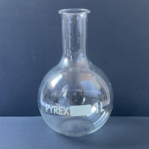 以下が含まれることがあります： 細い首を持つ透明なPyrex 1L丸底フラスコ。フラスコは丸みを帯びた底を持ち、透明なガラスでできています。「PYREX」の文字がフラスコの側面に印刷されています。