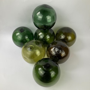 May include: A collection of seven spherical glass fishing floats arranged in a cluster. The floats vary in color from light olive green to deep forest green and dark amber, each featuring a small circular seal mark on the surface.