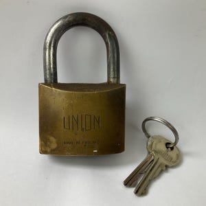 Puede incluir: Un candado de latón marca Union con un arco de acero desgastado y un juego de llaves en una anilla metálica. El cuerpo del candado tiene grabada la inscripción UNION y MADE IN ENGLAND, mostrando signos de desgaste y arañazos en la superficie.