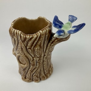Puede incluir: Un jarrón de cerámica con forma de tronco de árbol, con un pájaro azul posado en una rama. El jarrón es marrón con detalles de corteza texturizada. El pájaro tiene alas azules y pecho verde. Un objeto decorativo.