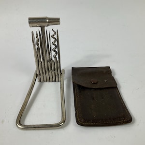 May include: A vintage multi-tool with a hammer, corkscrew, and blades, held in a metal frame. A dark brown leather case with a snap closure is next to the tool. The tool is made of stainless steel. The multi-tool is a useful item.