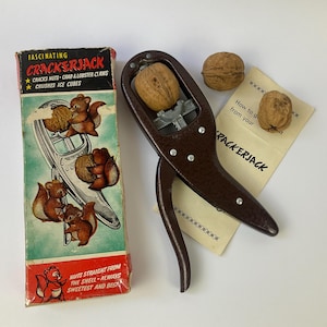 May include: A brown metal nutcracker with a silver-toned internal mechanism, accompanied by its original cardboard box featuring squirrel illustrations and the text CRACKERJACK, along with two whole walnuts and a printed instruction card.