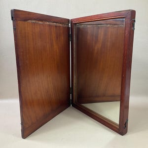 May include: A vintage, two-panel wooden vanity mirror. The mirror is framed in dark brown wood with a rectangular shape. The panels are connected by hinges, allowing them to open and close. The mirror is set against a neutral background.