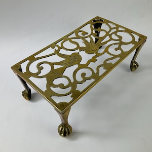 May include: A rectangular brass trivet featuring an openwork design of two birds surrounded by scrolling patterns. The piece is supported by four legs, each terminating in a claw-and-ball foot, providing a raised platform for kitchenware.