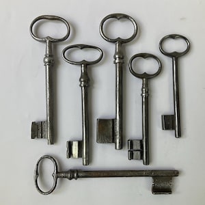 Puede incluir: Colección de llaves antiguas de metal de varios tamaños y diseños. Las llaves tienen mangos ornamentados y paletones rectangulares. Las llaves están dispuestas sobre una superficie blanca. Las llaves son de metal oscuro y envejecido.