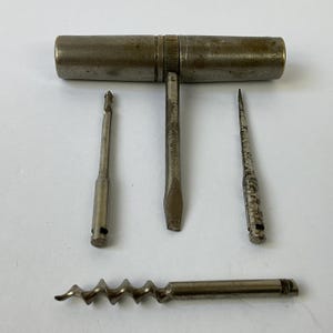 May include: A vintage multi-tool with a T-shaped handle and several interchangeable bits. The bits include a drill bit, a flathead screwdriver, a pointed awl, and a corkscrew. The tool is made of metal and shows signs of age and use.