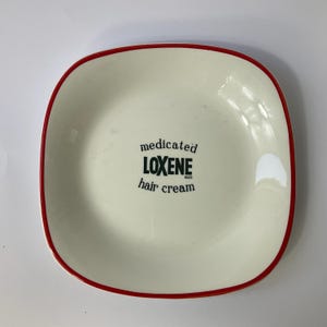Op de afbeelding: Een vierkant, crèmekleurig keramisch bord met een rode rand. Het bord heeft de tekst "medicated LOXENE hair cream" in zwart. Het bord heeft afgeronde hoeken en een licht verhoogde rand.