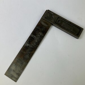 Op de afbeelding: Een donker metalen winkelhaak met een verweerde, geoxideerde afwerking op het blad. Het handvat heeft een verzonken ontwerp met een rond gat en de in reliëf aangebrachte tekst "JAMES CHESTERMAN" op het oppervlak van het metalen onderdeel.