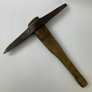 May include: A vintage pickaxe with a wooden handle and a dark, metallic head. The pickaxe head has a pointed end and a flat, chisel-like end. The wooden handle is a light brown color.