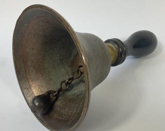 May include: A bronze-colored hand bell with a dark wooden handle. The bell has a rounded shape with a clapper attached by a chain. The handle is smooth and rounded, and the bell appears to be made of metal.