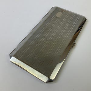May include: A rectangular, silver-toned cigarette case with a textured, vertically striped pattern on the lid. The case has a small, rectangular decorative element near the top and a clasp on the side. The edges are polished and reflective.