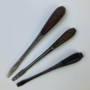 May include: Three vintage flathead screwdrivers with tapered metal shafts and rounded handles. The handles are made of dark brown, medium brown, and black material. Each tool features a flat, rectangular tip designed for slotted screws.