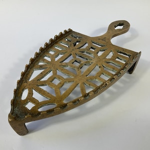 May include: A vintage brass iron rest with an openwork design. The iron rest has a handle and a curved base. The surface is decorated with a geometric pattern. The iron rest is designed to hold a hot iron.