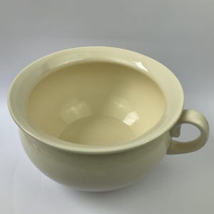 May include: A cream-colored chamber pot with a rounded body and a wide, flat rim. It has a single, curved handle on the side. The interior is a lighter shade of cream, and the overall design is simple and functional.