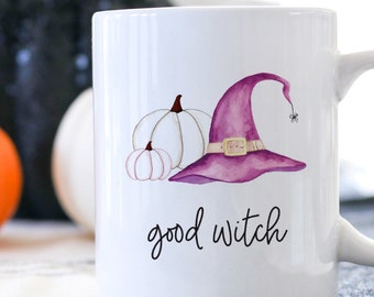 May include: White ceramic mug with a watercolor design of a purple witch hat, two white pumpkins, and the words "good witch" in black script. An orange pumpkin and other decorative items are in the background.