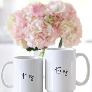 May include: Two white ceramic mugs with black handwritten text. The larger mug says "15 oz" and the smaller mug says "11 oz".