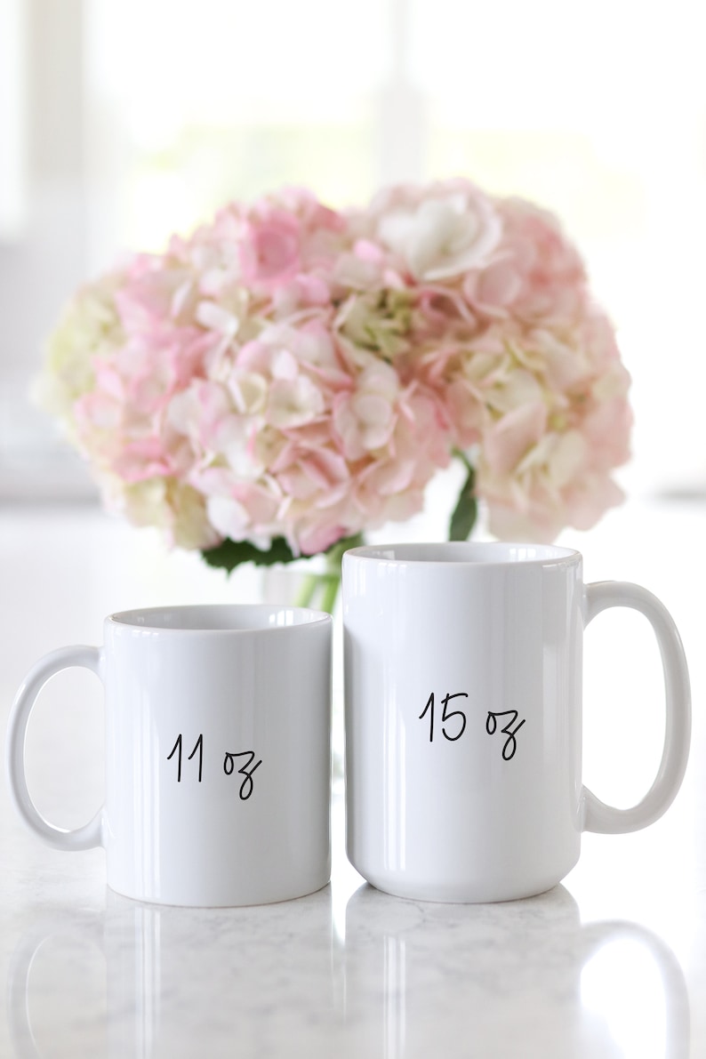 May include: Two white ceramic mugs with black handwritten text. The larger mug says "15 oz" and the smaller mug says "11 oz".