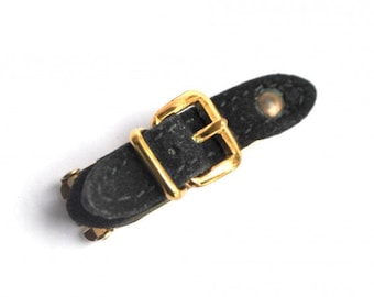 May include: A black leather strap with a gold buckle. The strap has a textured surface and a small gold stud.