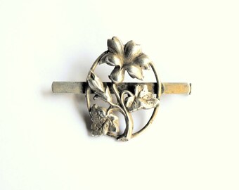 May include: Silver tone flower brooch with a circular design and a bar pin. The flower has five petals and is surrounded by leaves.