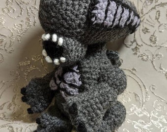 May include: A crocheted gray and purple alien plush toy with white teeth. The toy is sitting on a white patterned surface.