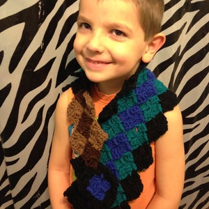 May include: A young boy wearing a colourful crocheted scarf with brown, blue and green squares. The scarf is tied around his neck and hangs down his chest. He is smiling and looking at the camera.