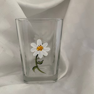 May include: A clear glass tumbler with a hand-painted white daisy with a yellow center. The daisy is painted on the side of the glass.