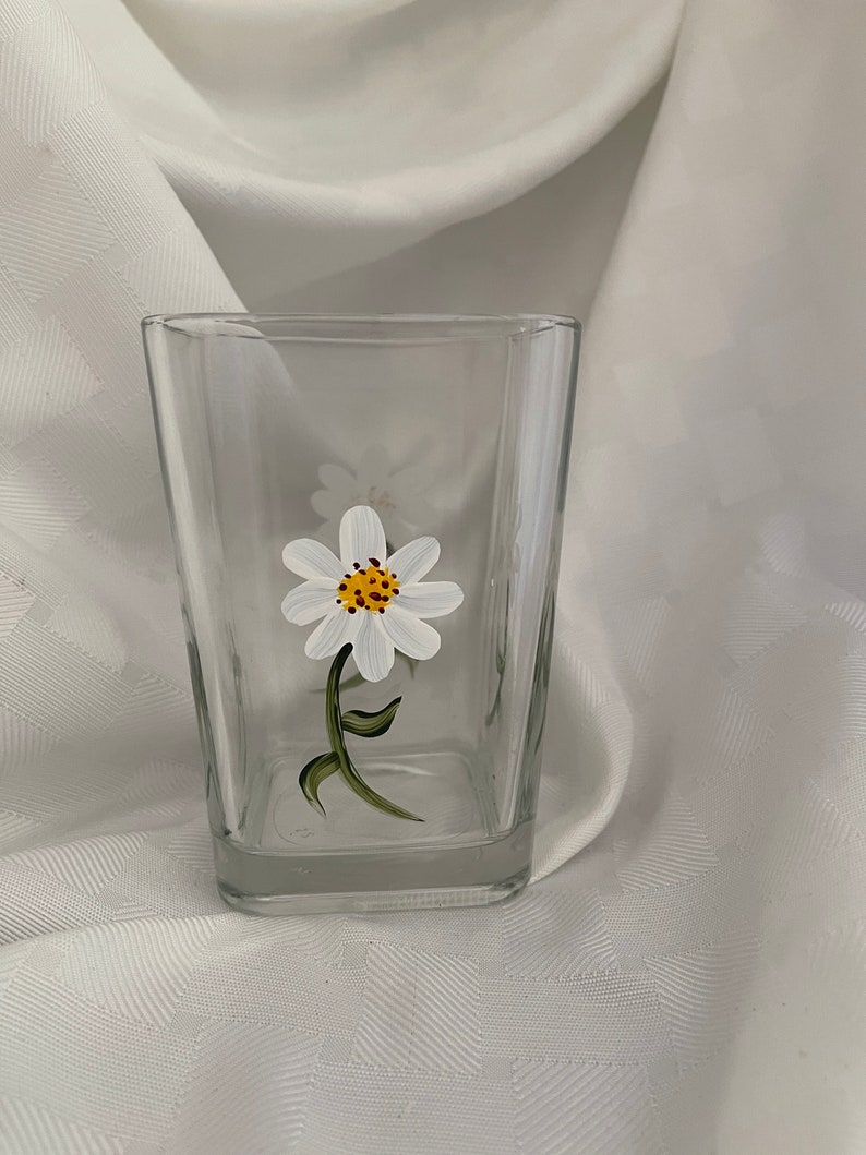 May include: A clear glass tumbler with a hand-painted white daisy with a yellow center. The daisy is painted on the side of the glass.