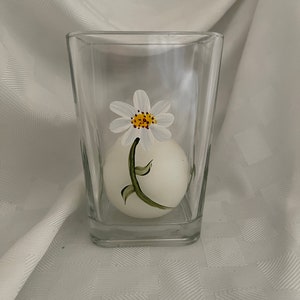May include: A clear glass candle holder with a white candle inside. The candle holder has a painted white daisy with a yellow center and green stem.