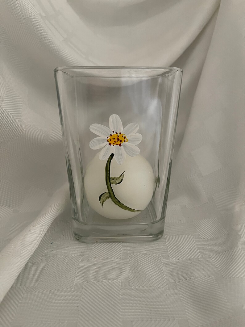 May include: A clear glass candle holder with a white candle inside. The candle holder has a painted white daisy with a yellow center and green stem.