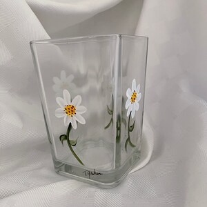 May include: A clear glass vase with a square shape, hand-painted with white daisies and green stems. The vase is signed "T. Graham" on the bottom.