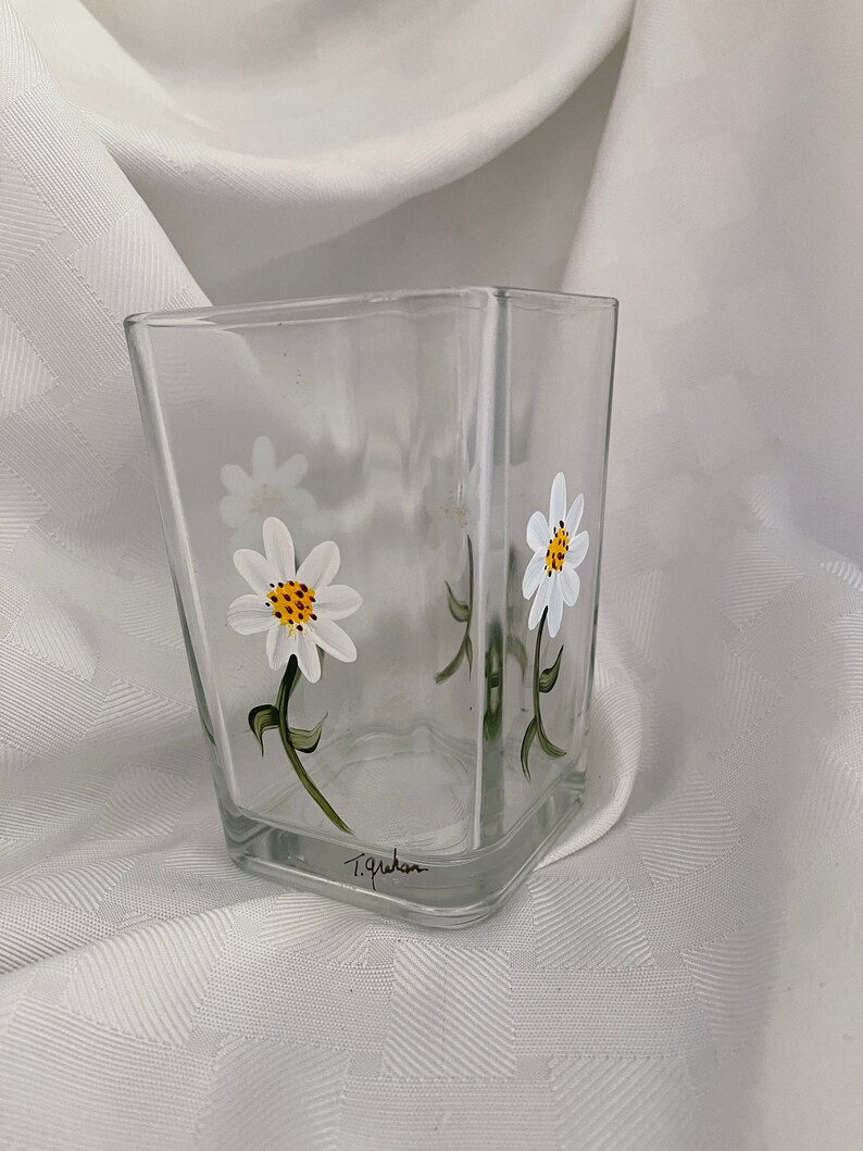 May include: A clear glass vase with a square shape, hand-painted with white daisies and green stems. The vase is signed "T. Graham" on the bottom.