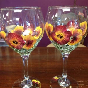 May include: Two clear wine glasses with hand-painted floral designs. The flowers are a mix of yellow, orange, and burgundy, with green accents. The stems and bases of the glasses are clear, and the glasses are set on a wooden surface.