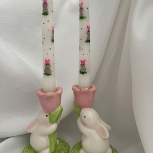May include: A pair of white ceramic bunny candle holders with pink flowers and green leaves. Each holder features a white taper candle with a pattern of gray bunnies and colorful dots.