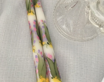 May include: Two white taper candles with hand-painted floral designs. The candles feature green leaves, pink and purple flowers, and yellow accents.