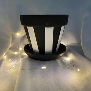 May include: Black and white striped ceramic flower pot with a matching saucer. The pot features alternating vertical black and white stripes. The pot is sitting on a white surface with small, warm white lights.