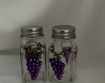 May include: A set of two glass salt and pepper shakers with silver lids. The shakers are decorated with a painted design of purple grapes on a vine.
