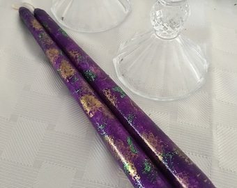 Peut inclure: Deux bougies violettes avec des accents de paillettes dorées et vertes. Les bougies sont sur une nappe blanche à motif diamant.