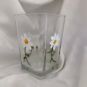 May include: A clear glass square vase with a hand-painted design of two white daisies with yellow centers and green stems.