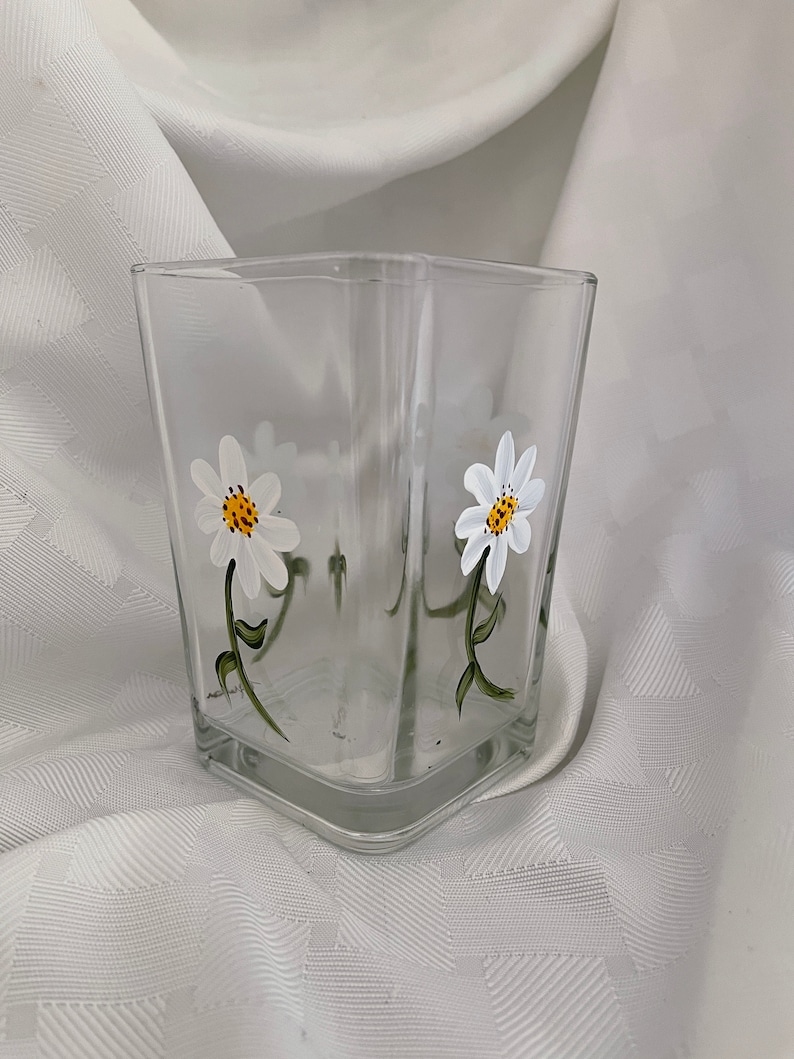 May include: A clear glass square vase with a hand-painted design of two white daisies with yellow centers and green stems.
