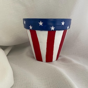 May include: A tapered flower pot featuring a hand-painted design with a dark blue rim adorned with white stars. The body of the pot displays vertical red and white stripes, creating a pattern reminiscent of the American flag design.