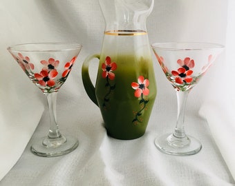 Könnte beinhalten: Ein Set aus drei grünen Glasgetränken mit handbemalten roten Blumen. Das Set enthält eine Karaffe und zwei Martinigläser.