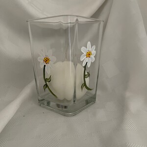 May include: A clear glass square vase with a white candle inside and hand-painted white daisies with yellow centers and green stems.