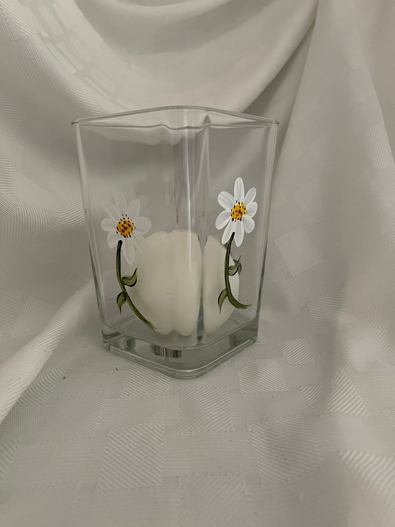 May include: A clear glass square vase with a white candle inside and hand-painted white daisies with yellow centers and green stems.