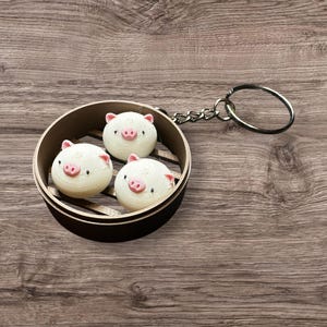May include: A white and pink pig-shaped keychain charm in a small wooden steamer basket with a silver chain and ring.